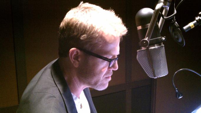 Scott Brick as seen from the side reading a book into a large microphone.  The recording studio is fairly dark, but he has a bright light shining on the text he is reading from.